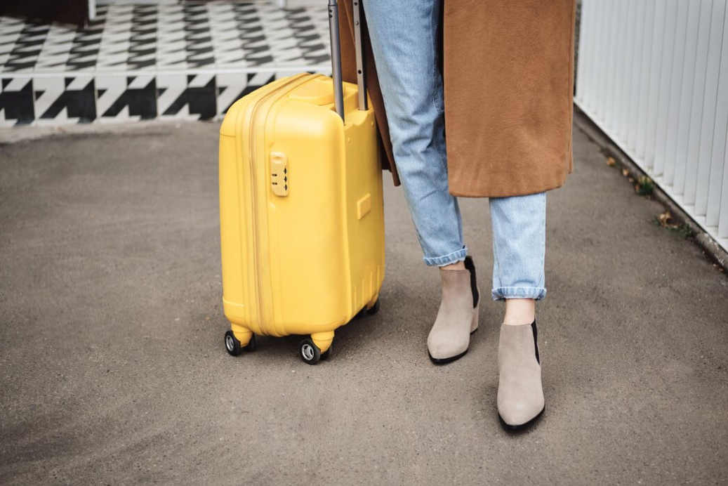 A person with a brown coat stands near a yellow suitcase on a patterned pavement